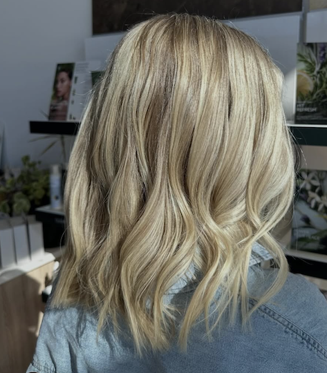 Blonde woman with wavy shoulder-length hair sitting in a salon or store, facing away from the camera.