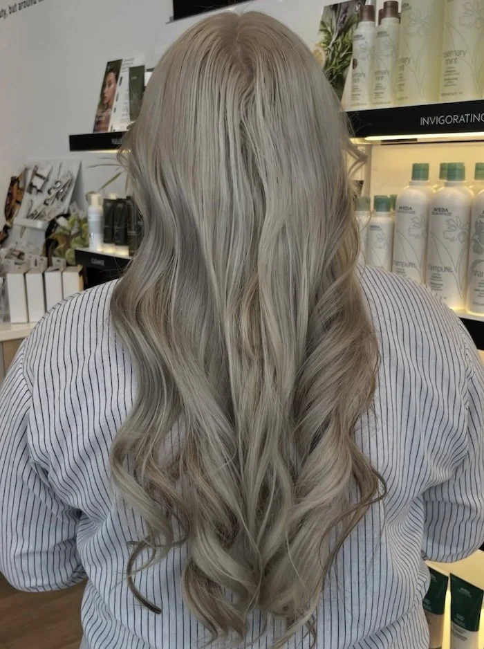 Back view of a woman with long, wavy, bleach-blonde hair in a retail store or salon, with shelves of hair care products in the background.