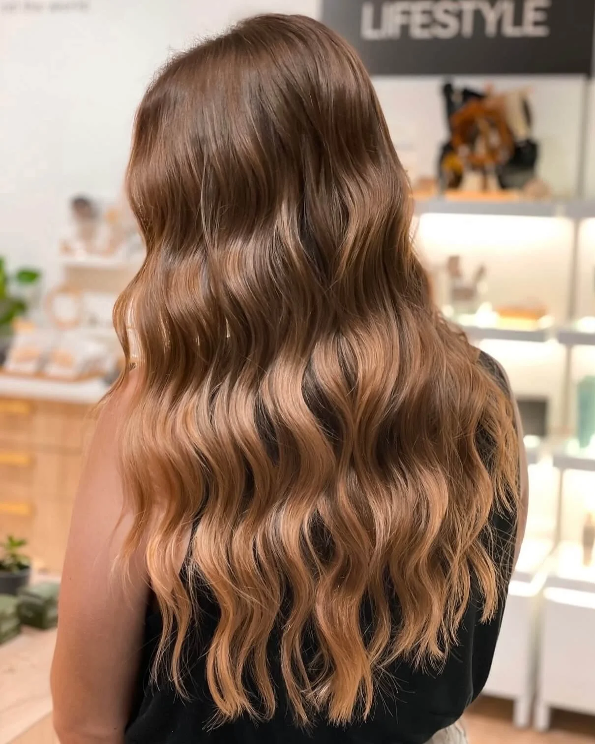 Back view of a woman with long, wavy, ombré hair transitioning from brown at the roots to blonde at the ends, standing in a salon.