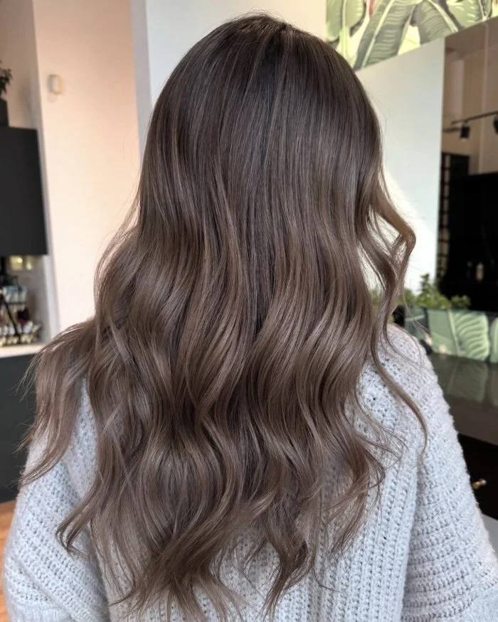 Back view of a woman with long, wavy, light brown hair, wearing a knitted gray sweater, in a modern salon.