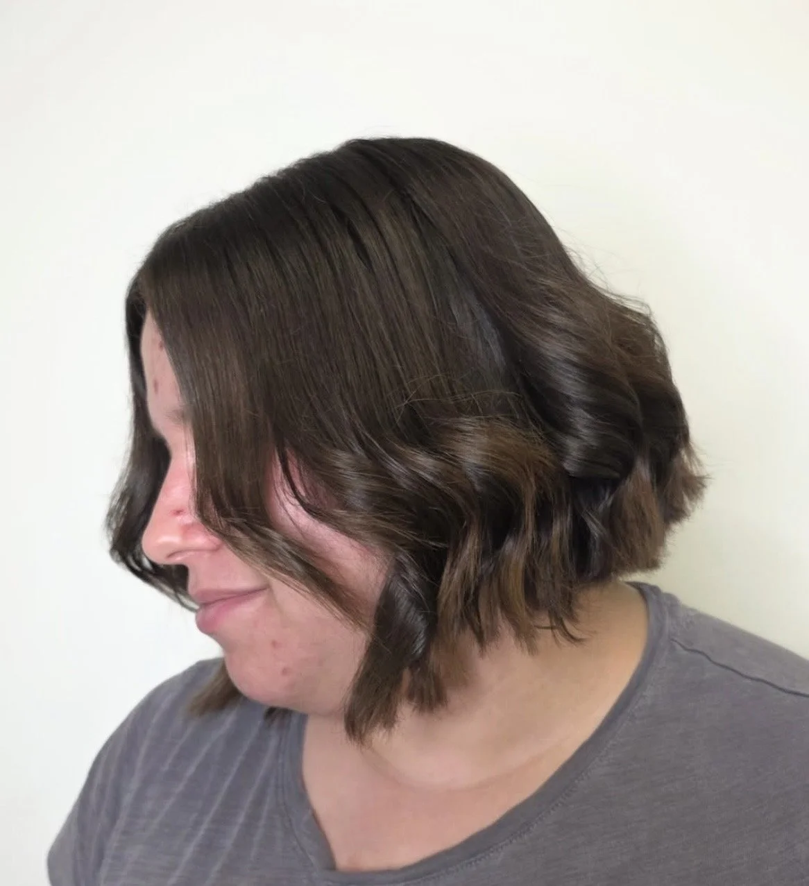 Side view of a woman with shoulder-length brown hair styled in loose, wavy curls, wearing a gray shirt, against a plain white background.