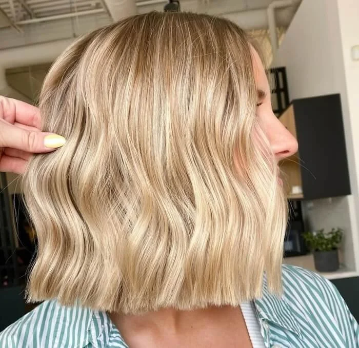 A woman with shoulder-length blonde wavy hair is getting her hair styled, with a hand holding a section of her hair and a salon interior in the background.