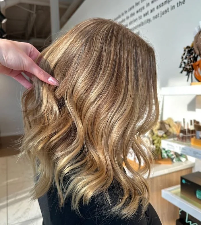 Close-up of a woman with shoulder-length, wavy blonde hair with darker roots, held by a hand pinching a small section of hair.