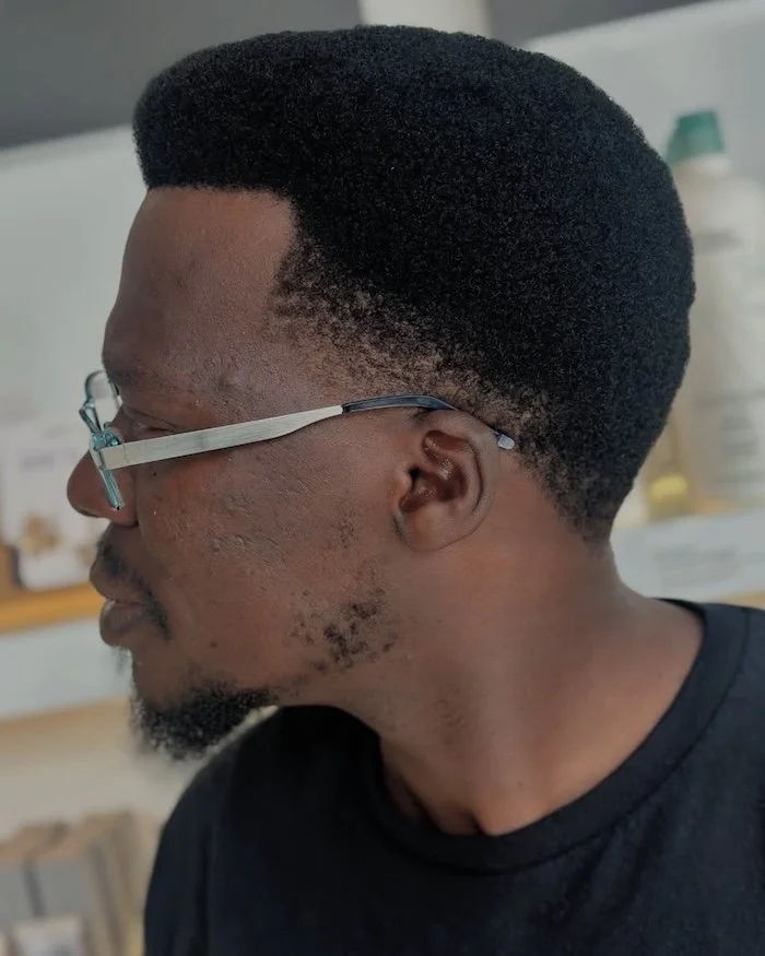 Side profile of a man with short, textured black hair, wearing eyeglasses, in a room with a blurred background.