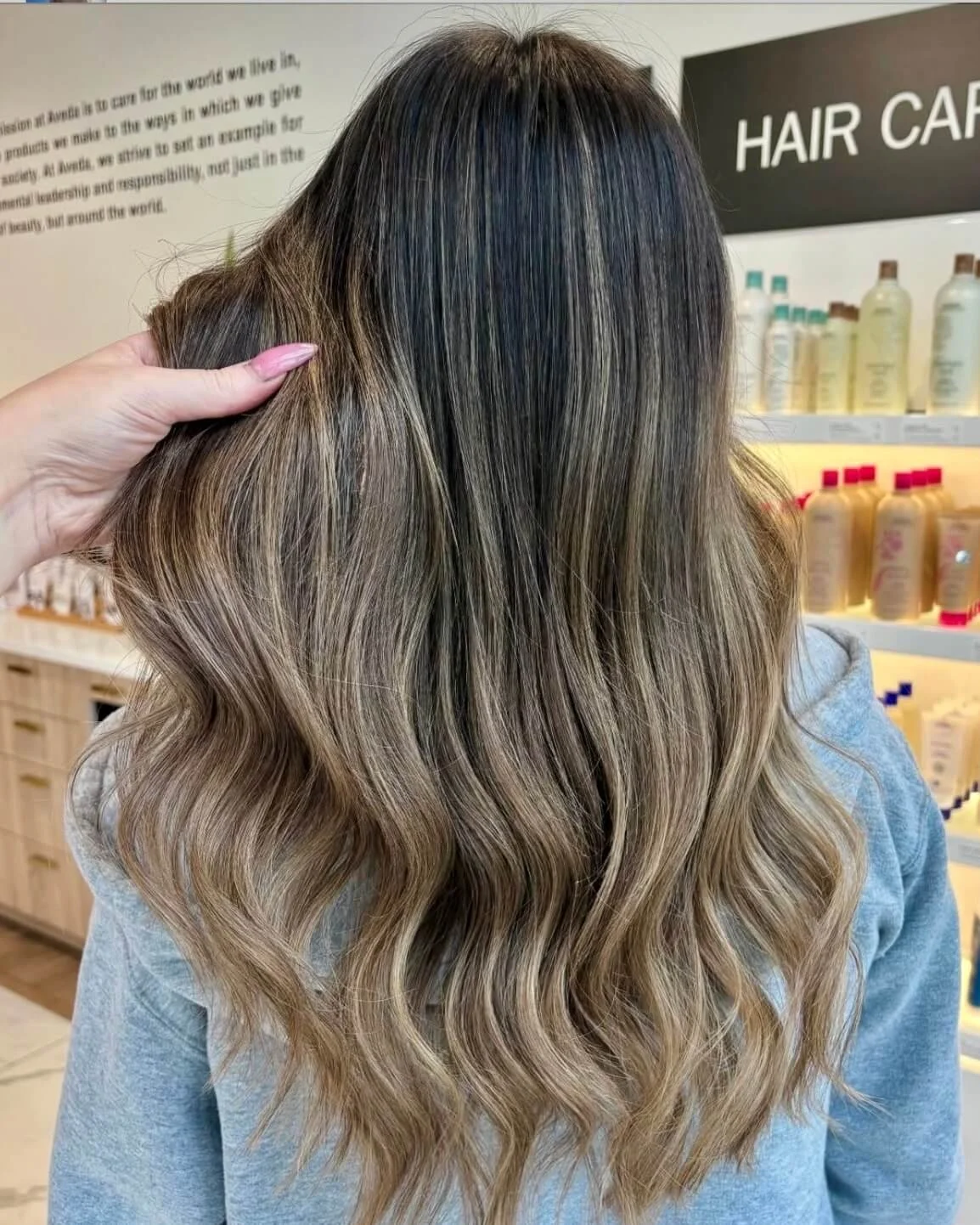 Back of a woman with long, wavy, ombre hair in shades of brown and blonde, at a hair care salon.