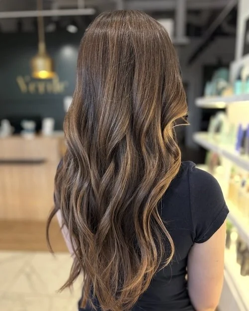 Back of a woman with long, wavy, light brown hair with highlights, wearing a black shirt, in a store or salon setting.