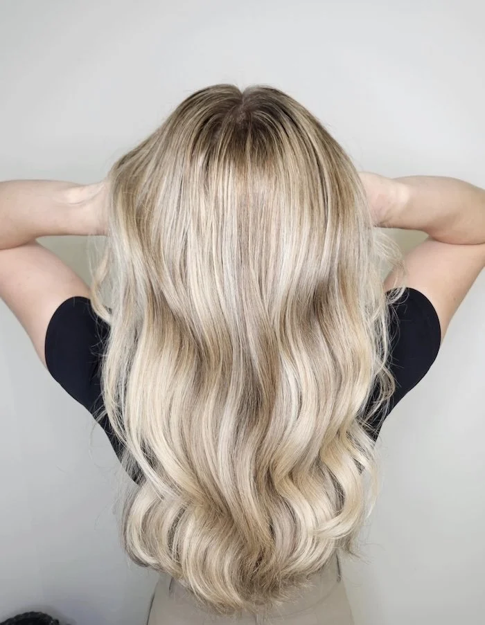 Back of a woman with long, wavy, blonde hair. She is wearing a black shirt and standing against a plain gray background.