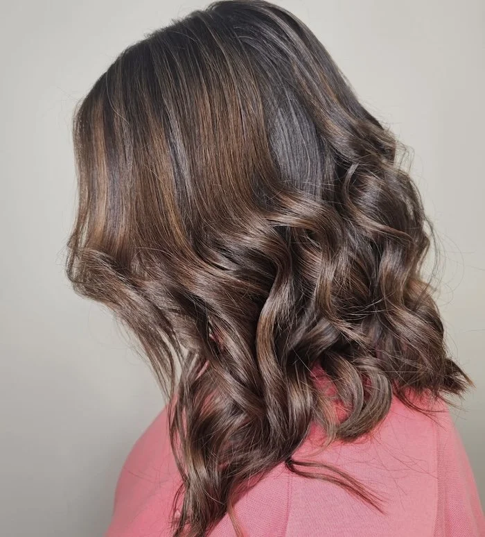 Close-up side view of a woman with wavy brown hair with highlights, wearing a pink top.
