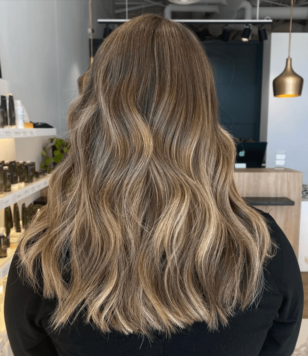 Back view of a woman with long, wavy, light brown hair with blonde highlights inside a salon.