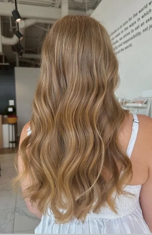 Back of a woman with long, wavy blonde hair, wearing a white spaghetti strap top, in a modern interior space.
