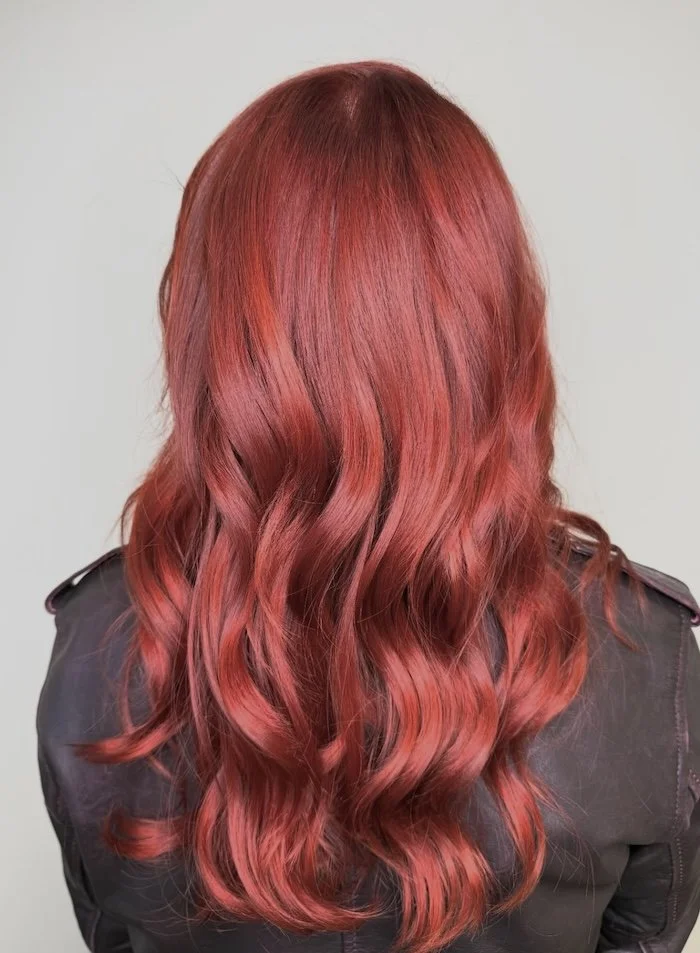 Back view of a person with long wavy red hair wearing a black leather jacket against a plain background.