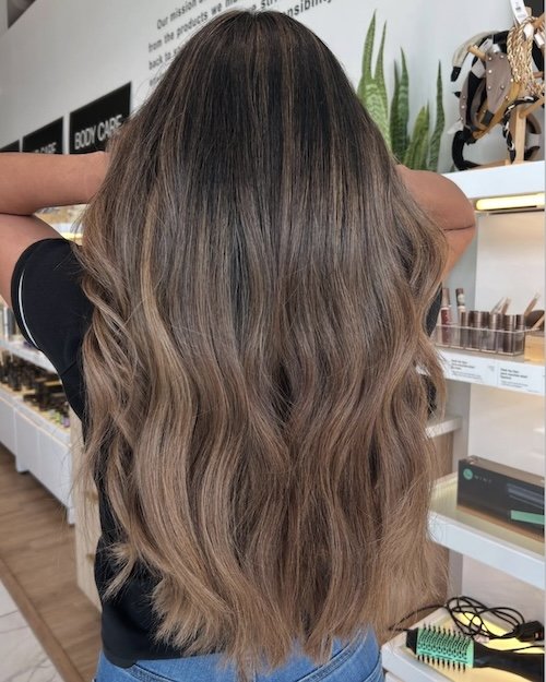 Back view of a woman with long, wavy, brown hair in a store, with beauty products and a plant in the background.
