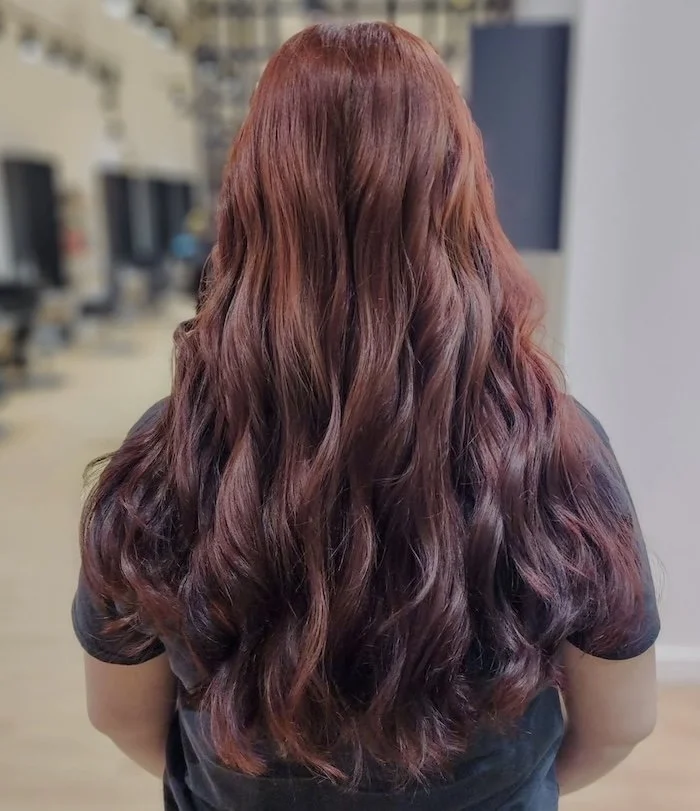 Back view of a woman with long, wavy, reddish-brown hair in a salon or indoor setting.