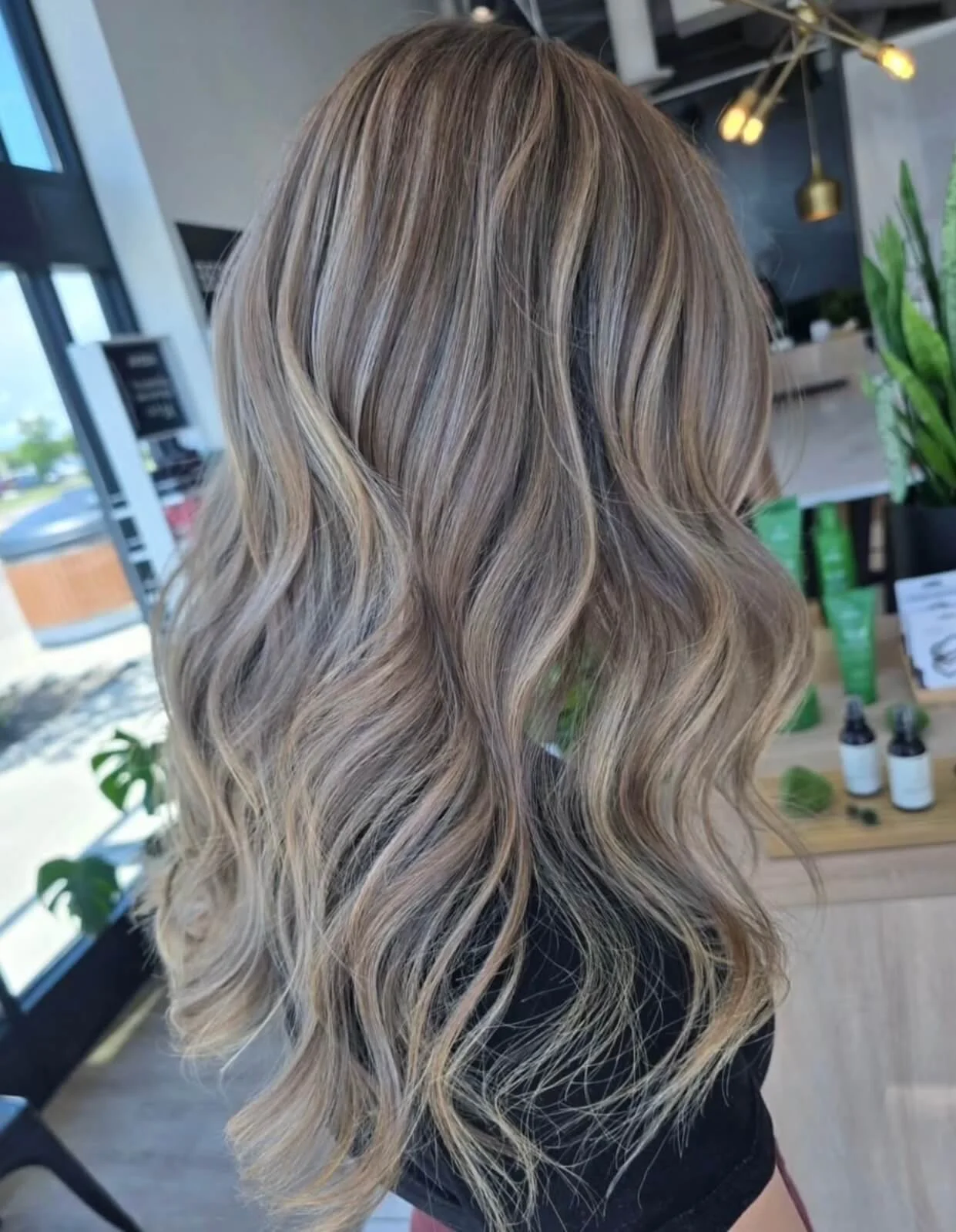 Back view of a woman with long, wavy blonde hair with darker roots, inside a modern salon.