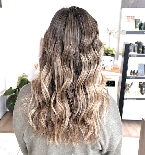 Back view of a woman with long, wavy, ombre hair in shades of blonde and light brown in a salon.