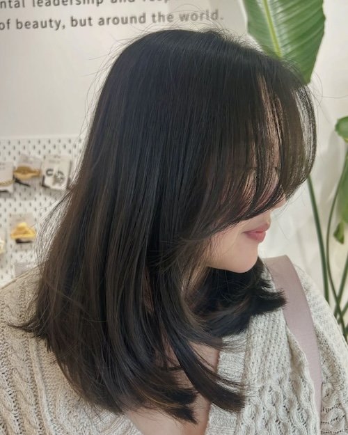 Side profile of a woman with shoulder-length dark brown hair and bangs, smiling slightly, in an indoor setting with a plant and a wall with text.