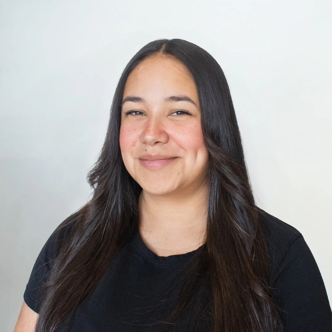 A young woman with long dark hair and a slight smile, wearing a black shirt, standing against a plain white background.