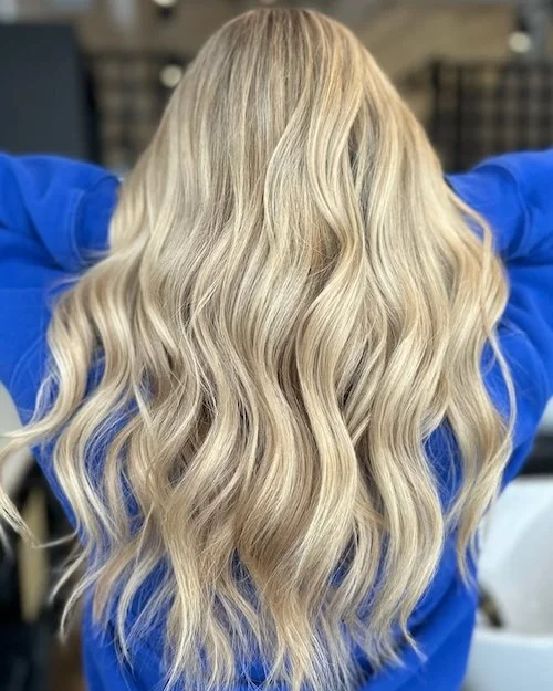 Back view of a woman with long, wavy blonde hair wearing a blue top.