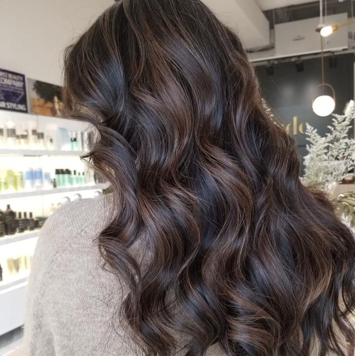 Close-up of wavy brunette hair with highlights, inside a salon with shelves of hair products in the background.