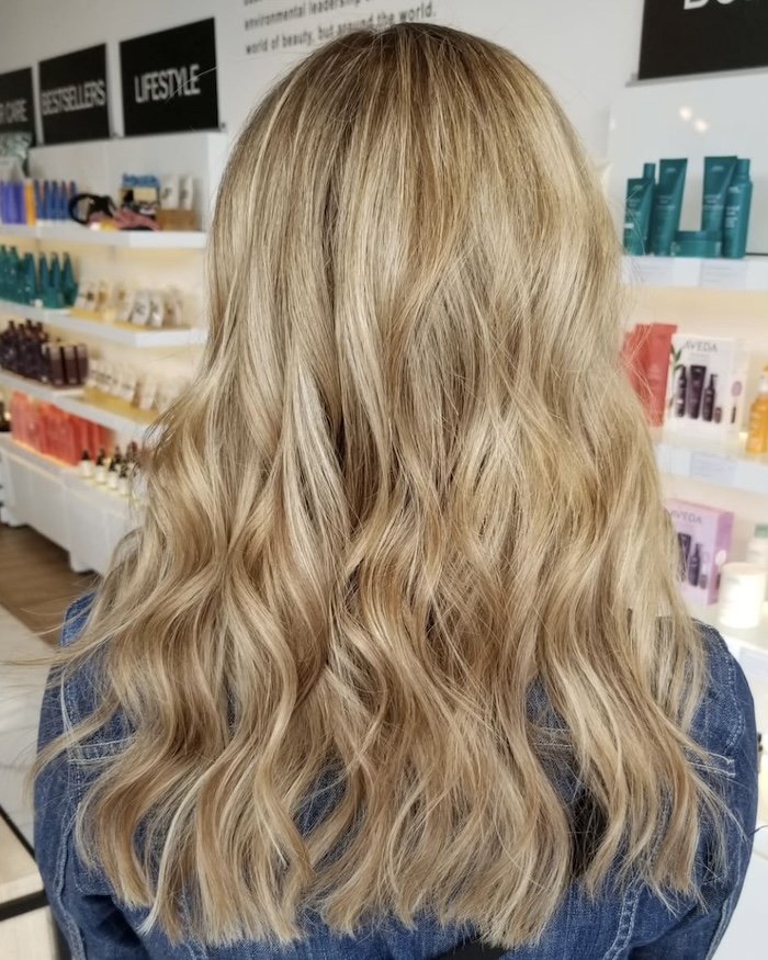 Back view of a person with long, wavy blonde hair in a store or beauty salon, with shelves of haircare products visible in the background.