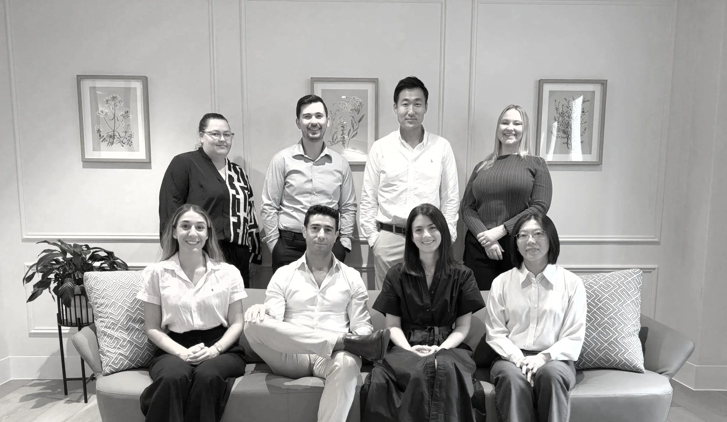 Group of nine diverse professionals, five women and four men, posing in an office or conference room with framed botanical paintings on the wall.