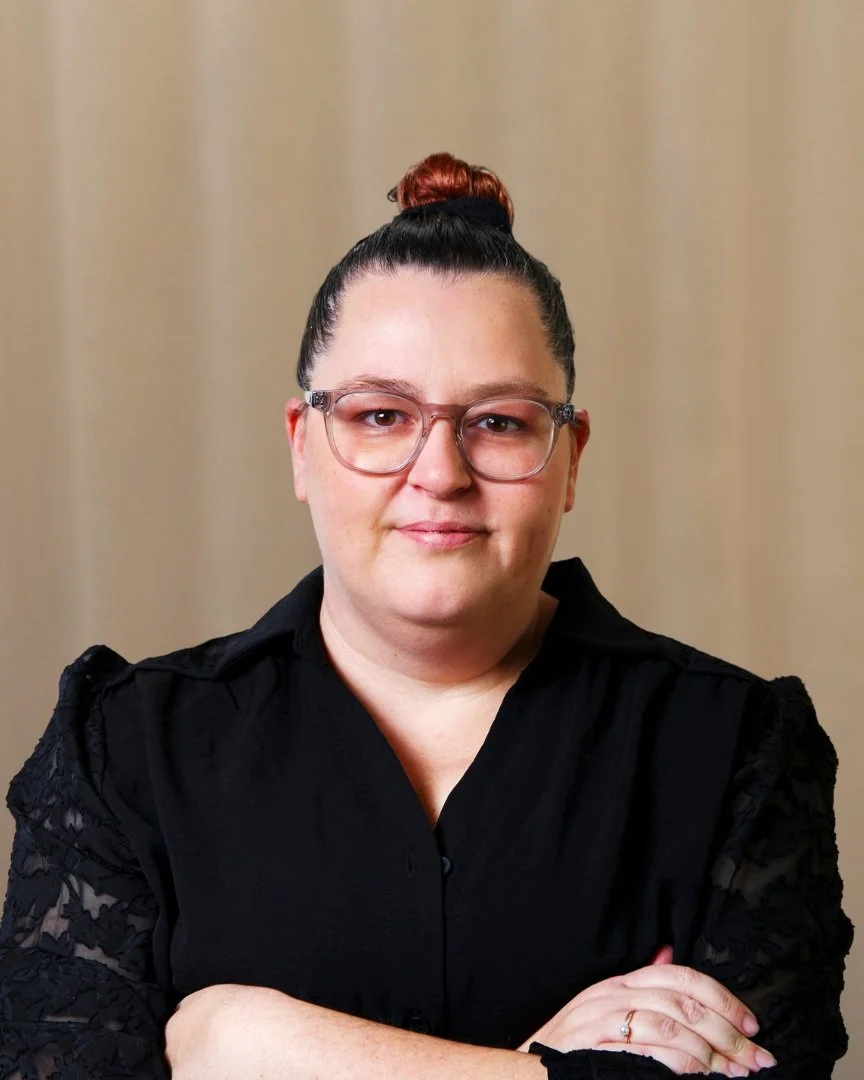 A woman with glasses, wearing a black blouse, with her arms crossed, standing in front of a plain beige background.