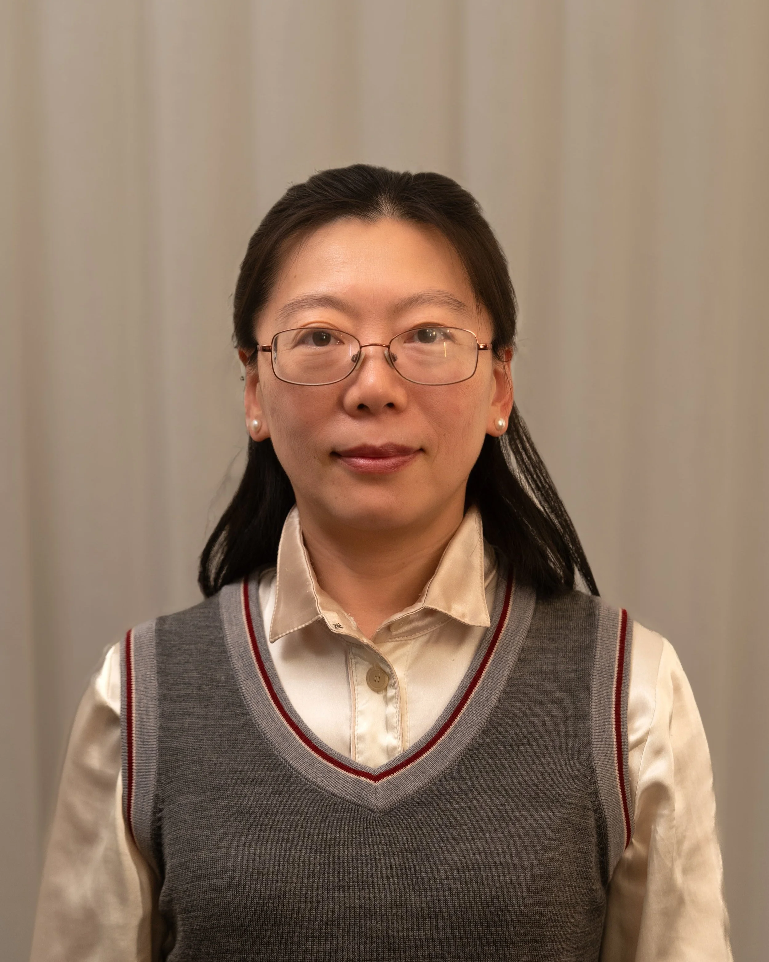 A woman with dark hair and glasses, wearing a beige shirt and a gray vest with red and white stripes, standing in front of a neutral background.