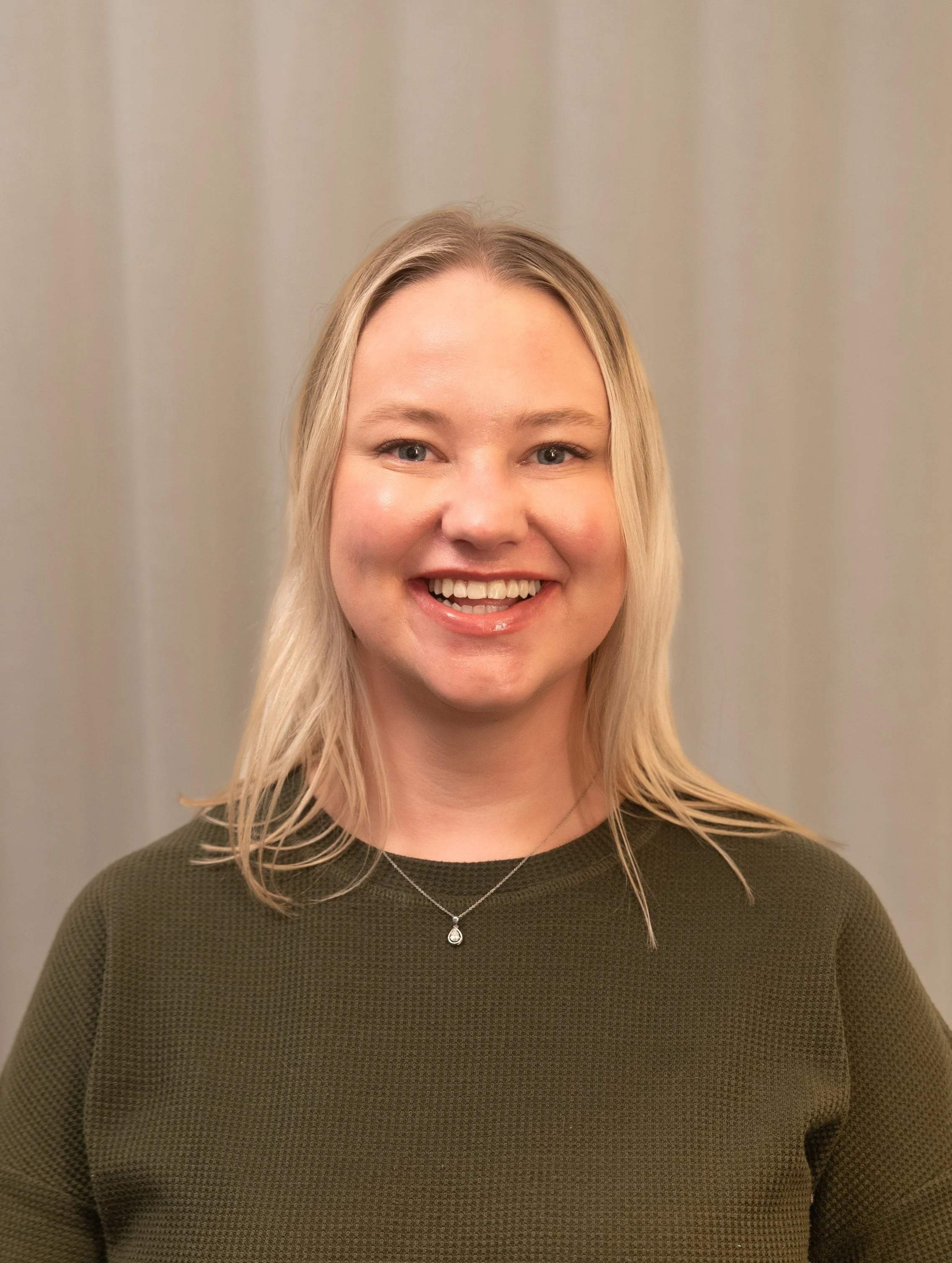 A woman with blonde hair smiling, wearing a dark green textured top and a delicate silver necklace with a teardrop pendant, standing against a neutral background.