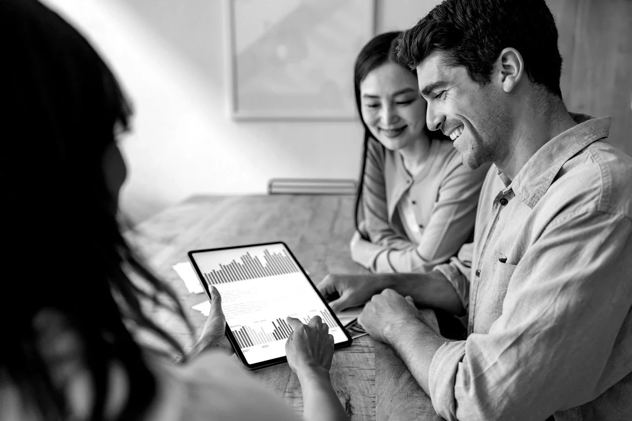 Three people sitting at a table, looking at a tablet displaying graphs and charts, smiling and engaged in a discussion.