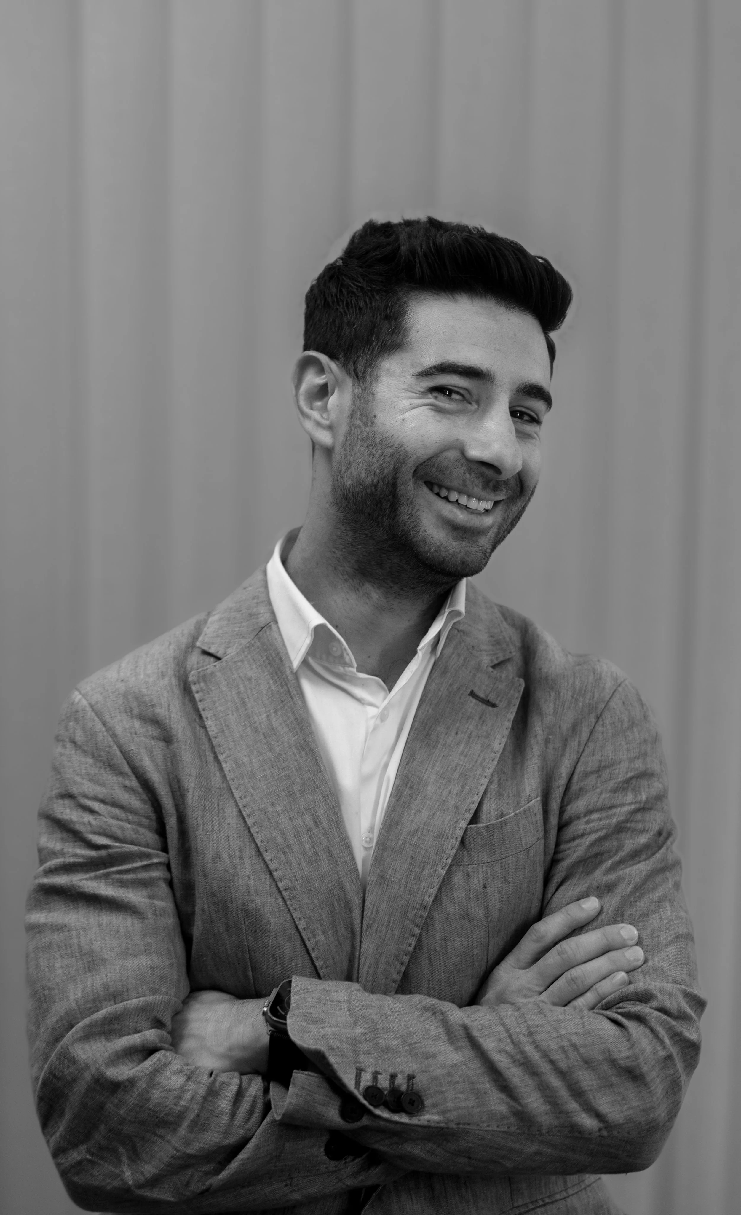 Black and white photo of a smiling man with dark hair, dressed in a light-colored suit and white shirt, standing with crossed arms in front of a simple wall background.