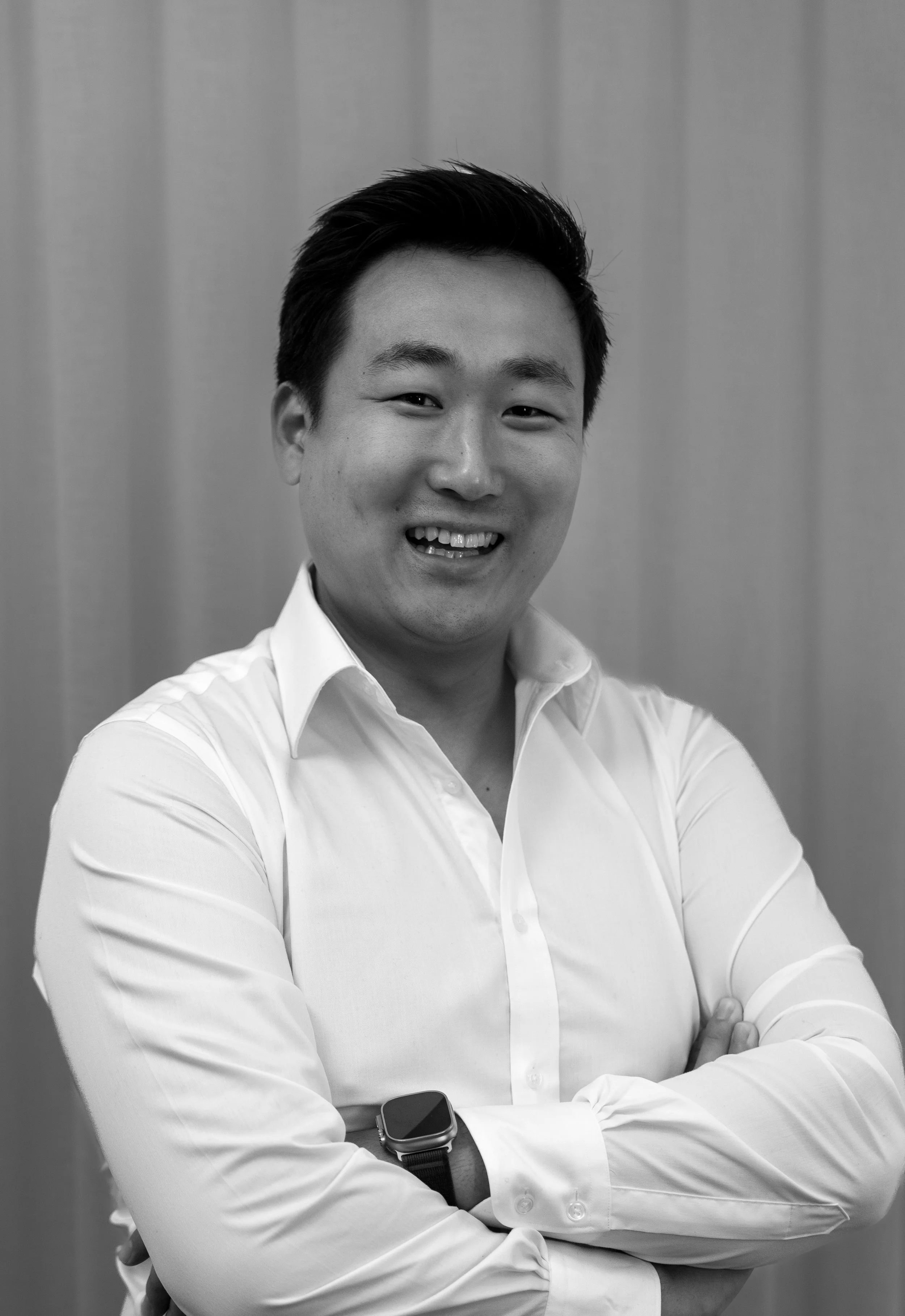 A Black-and-white photo of a smiling Asian man with short dark hair, wearing a white dress shirt with the sleeves rolled up, standing with arms crossed and a smartwatch on his left wrist, in front of a plain background.