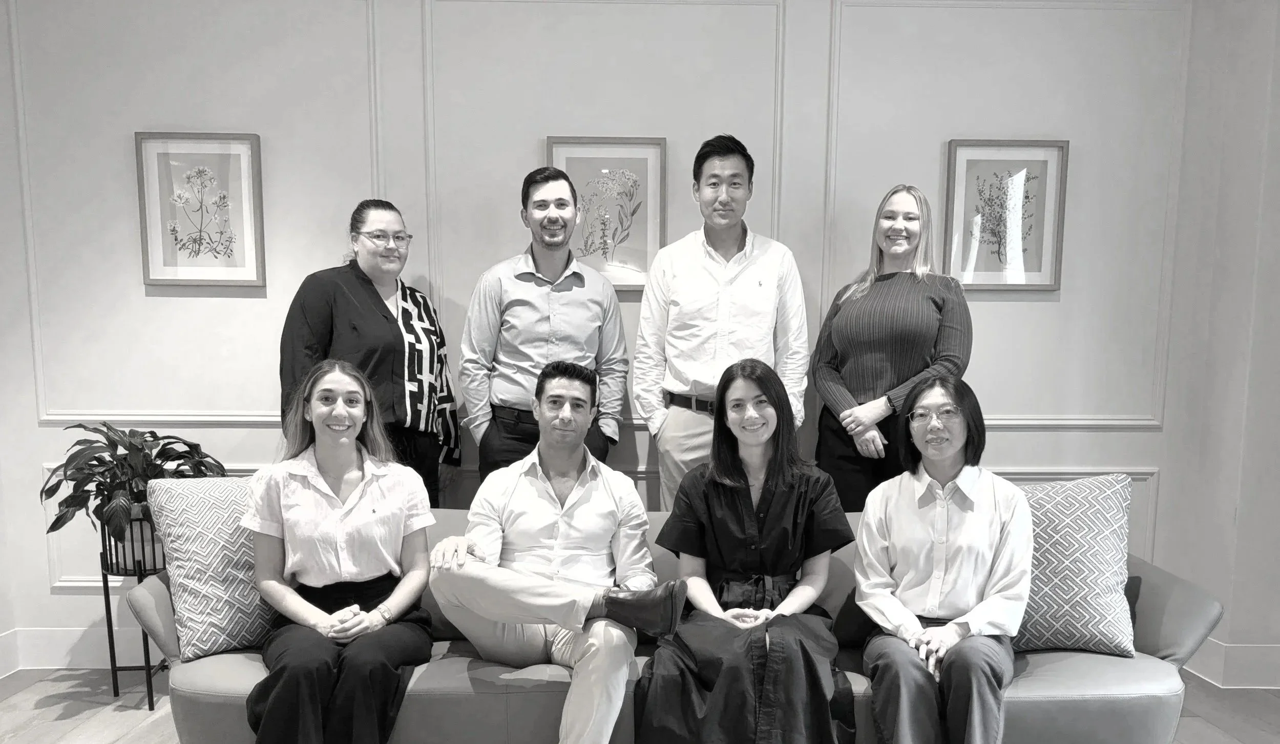 Group photo of nine people, four men and five women, in a well-lit room with framed botanical artwork on the wall, sitting on and standing around a sofa with patterned pillows.