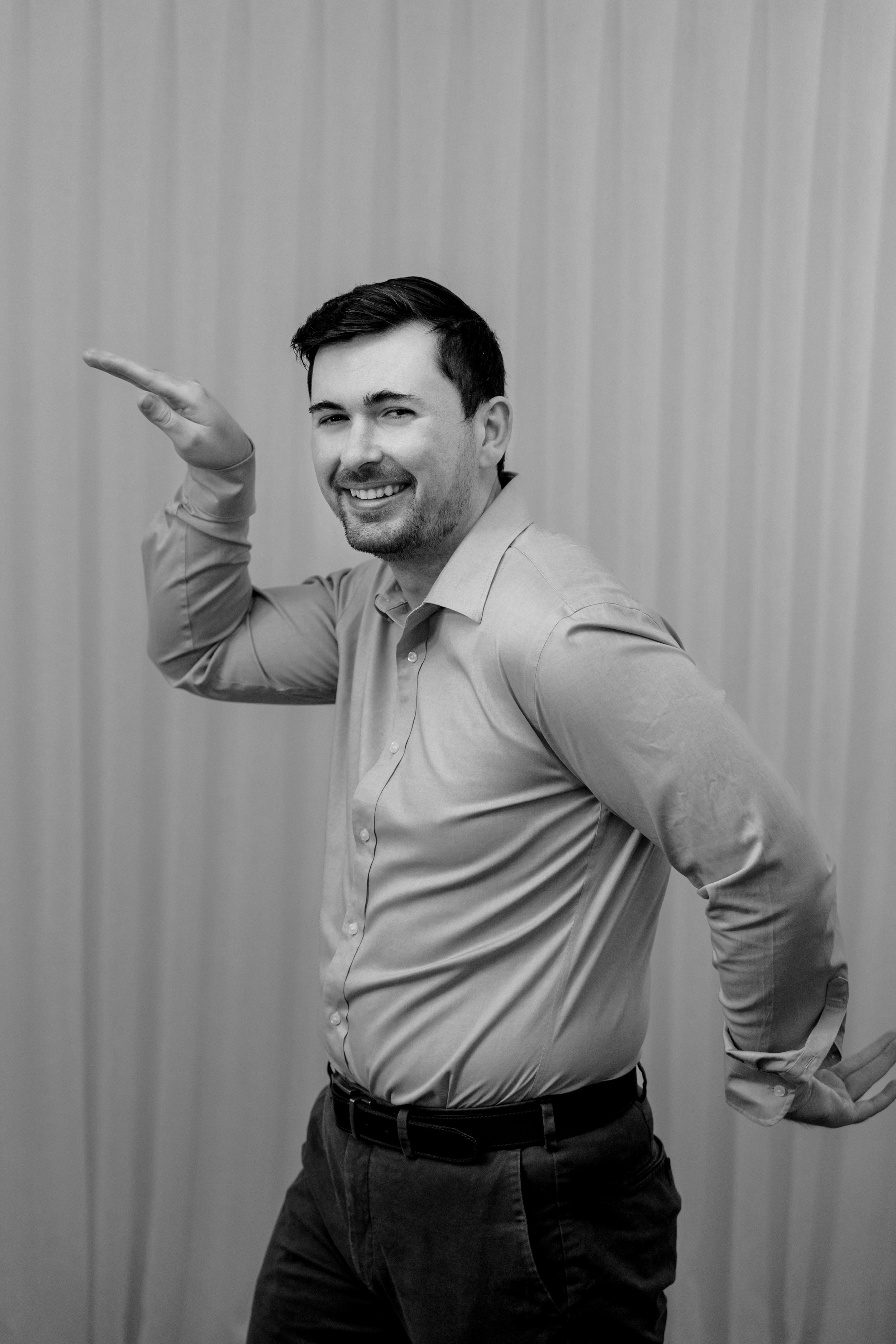 A black and white photo of a smiling man in a button-up shirt, with one arm raised and bent at the elbow, standing against a plain wall.