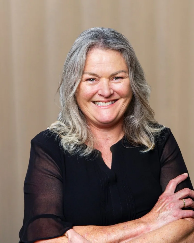 A smiling woman with gray hair and a black top standing against a beige background.