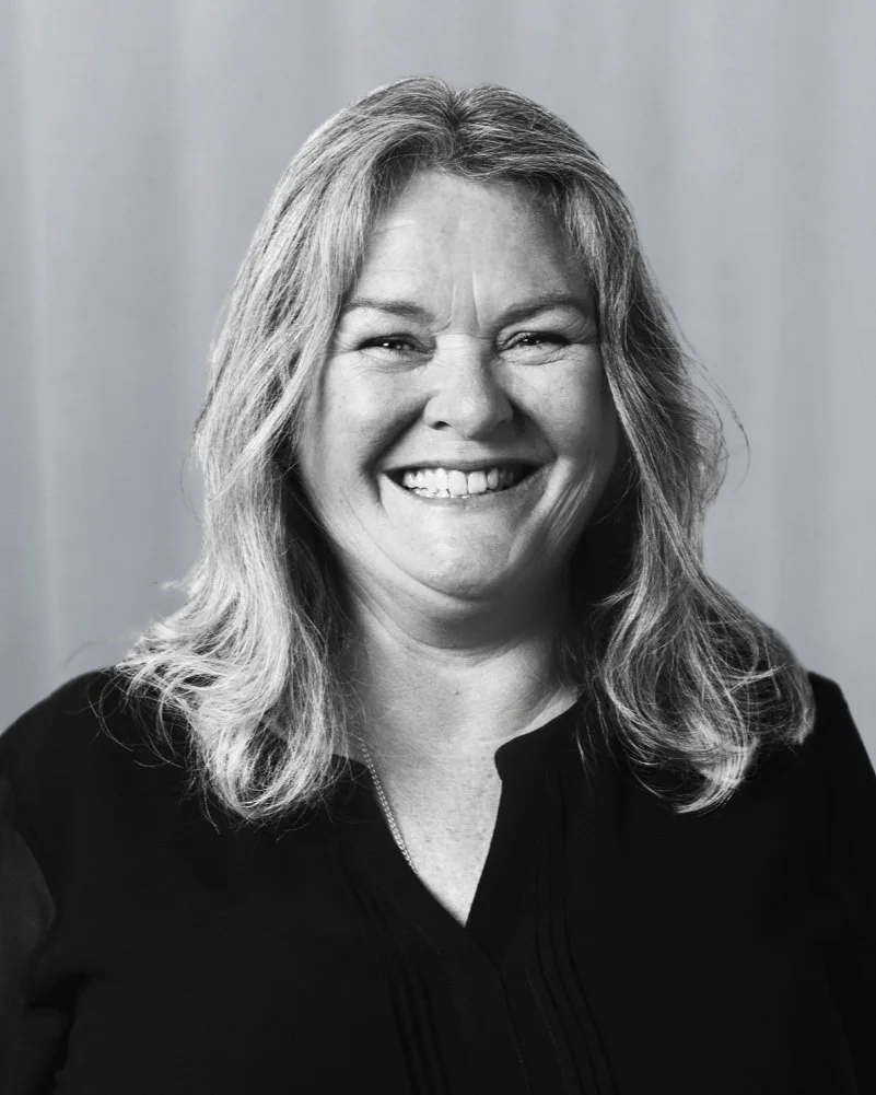 Black and white photo of a woman with shoulder-length wavy hair, smiling and wearing a dark top with a collar, against a plain background.