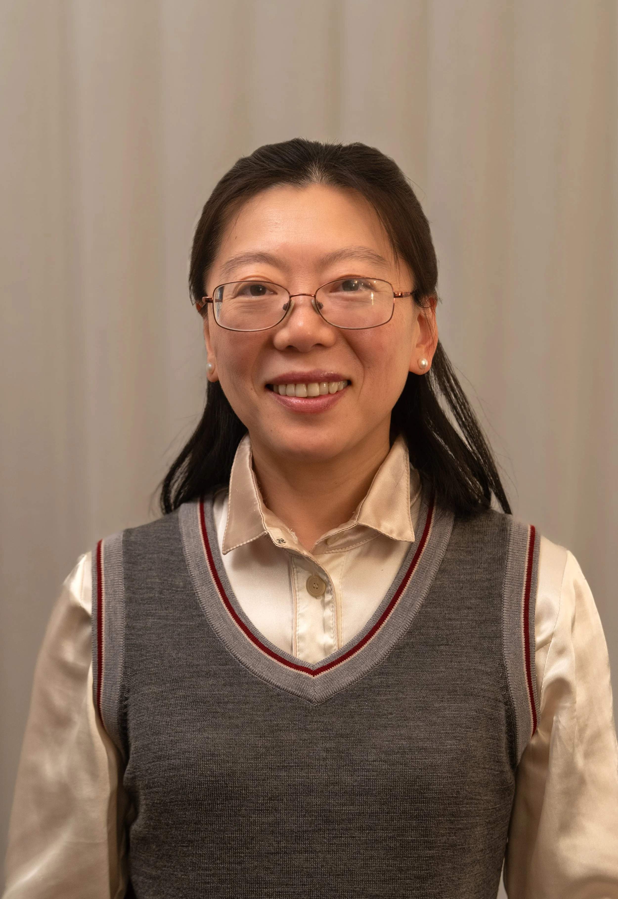 A woman with glasses and earrings, wearing a beige shirt underneath a gray vest with red and white stripes, smiling and standing against a neutral background.