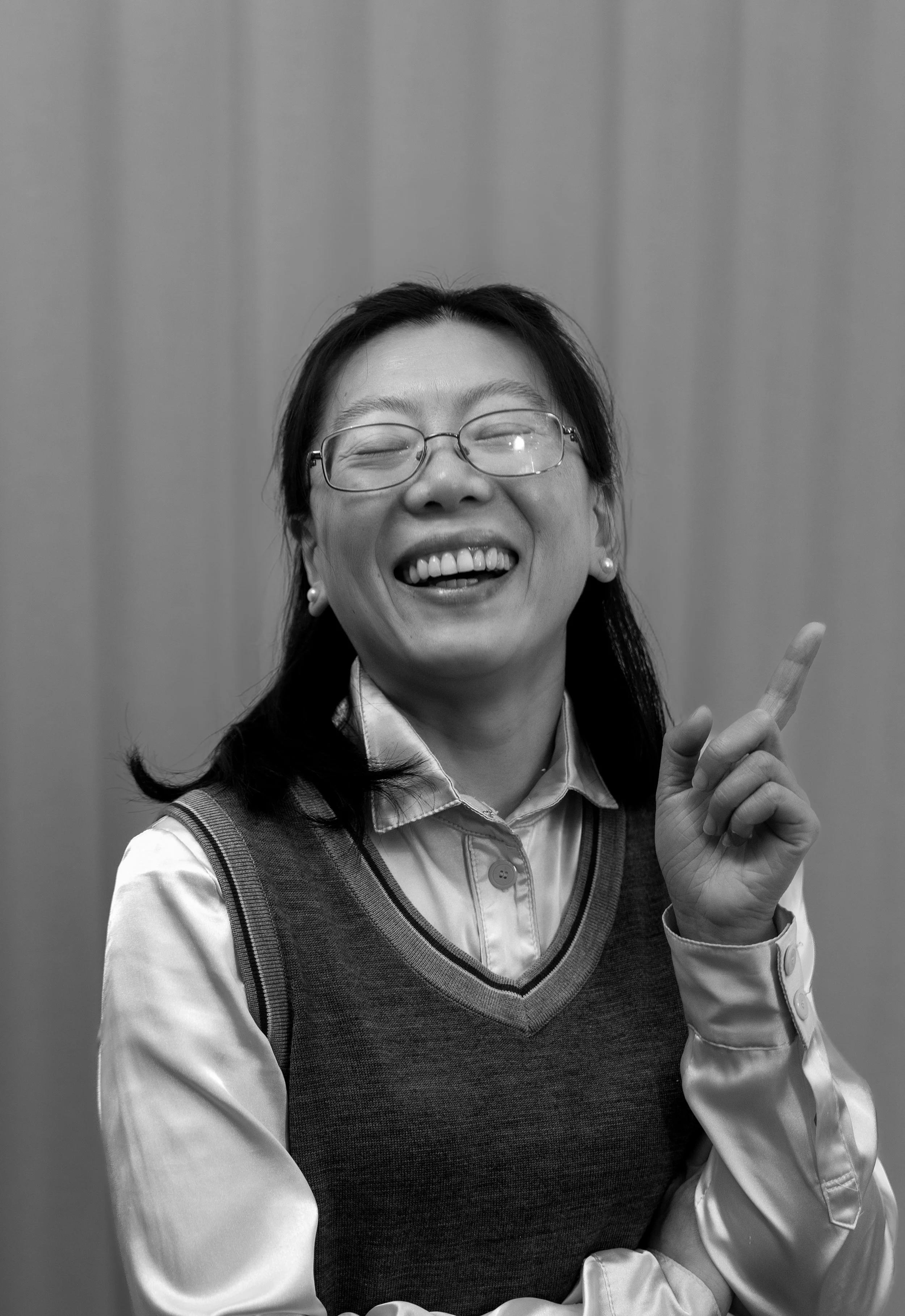 Smiling woman with glasses, wearing a sweater vest over a collared shirt, making a peace sign with her right hand in front of a plain wall.