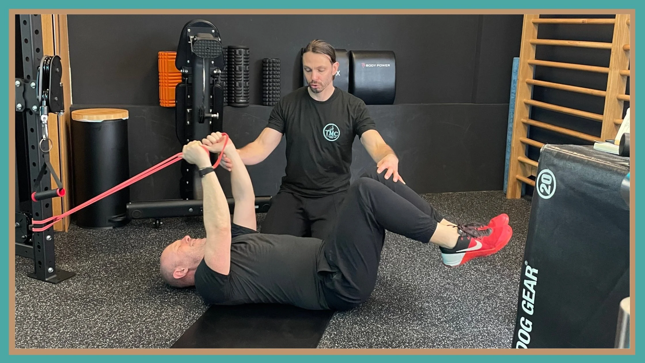 Personal trainer assisting a man with a resistance band exercise on a workout mat in a gym.