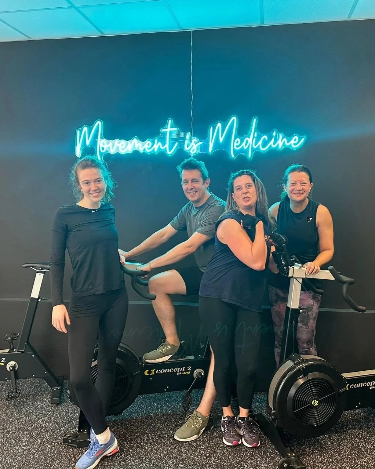 Four people in workout clothes posing on a stationary exercise bike in a gym with a neon sign that reads 'Movement is Medicine' on the wall behind them.