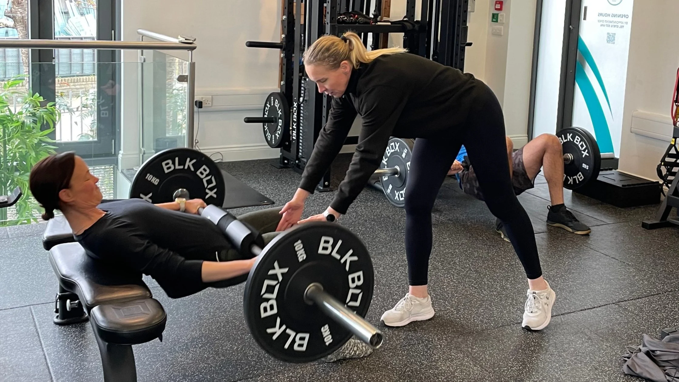 A woman lying on a workout bench performing a barbell exercises with a trainer assisting. The trainer stands next to her, reaching out to help. A man in the background is doing a leg lift exercise on the floor. The gym has weights and equipment around, natural light entering through windows, and a plant near the window.