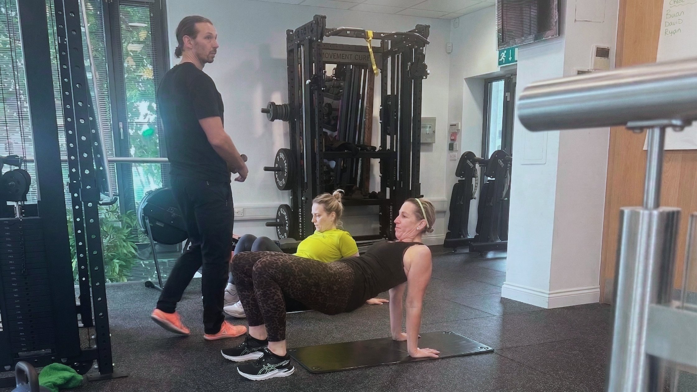 Three women exercising in a gym, with one in a yoga pose on a mat, another in a seated stretch, and a man standing nearby, surrounded by gym equipment.