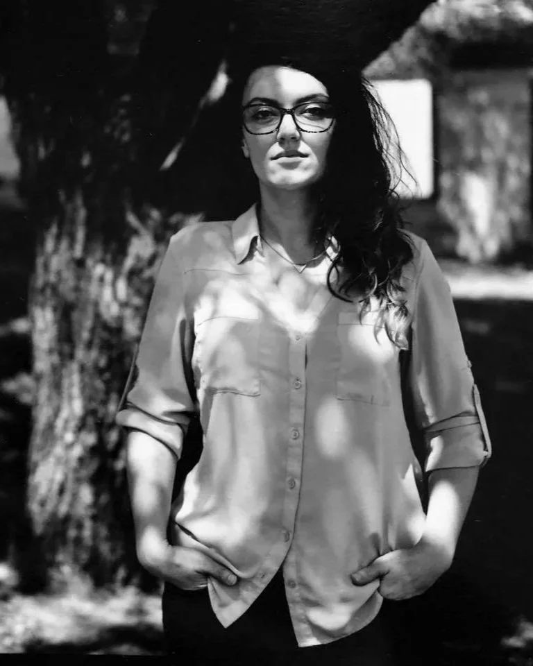 Black and white headshot of Kristina Smith, standing in front of tree, with long, wavy hair, and glasses. Image by Cameron Roe.