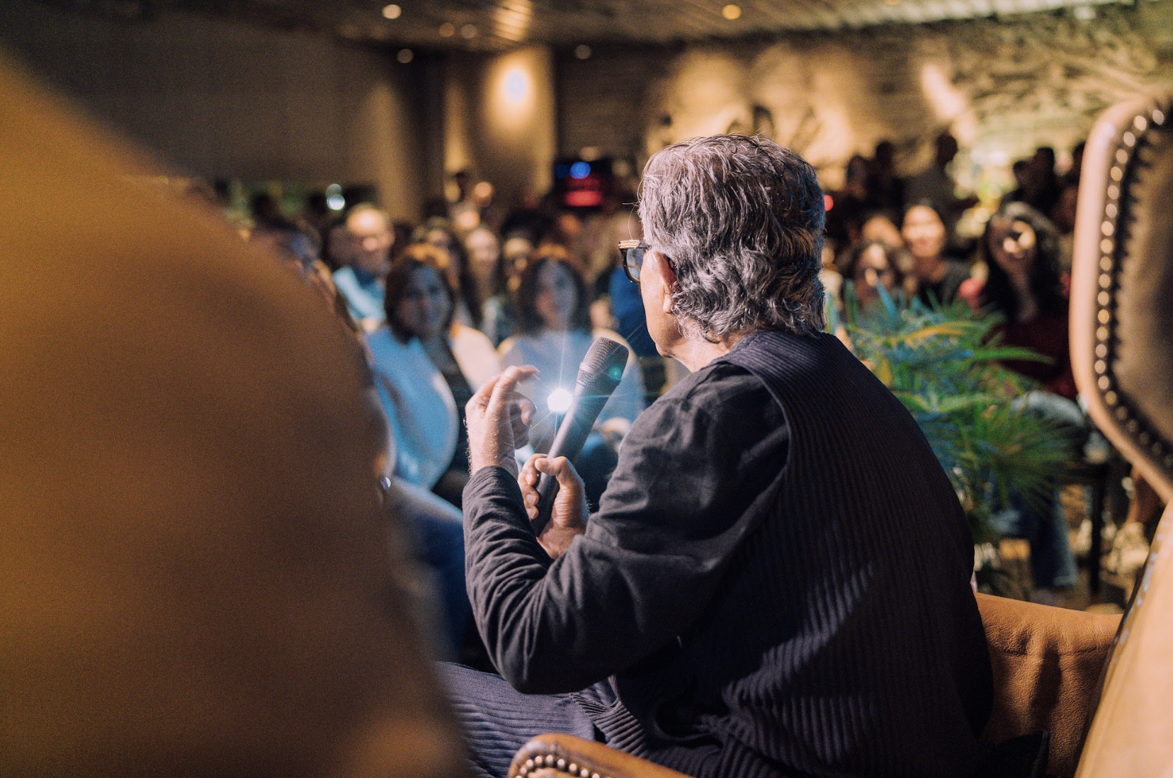 An elderly man with gray hair and glasses speaking into a microphone at a crowded indoor event with an audience in the background.