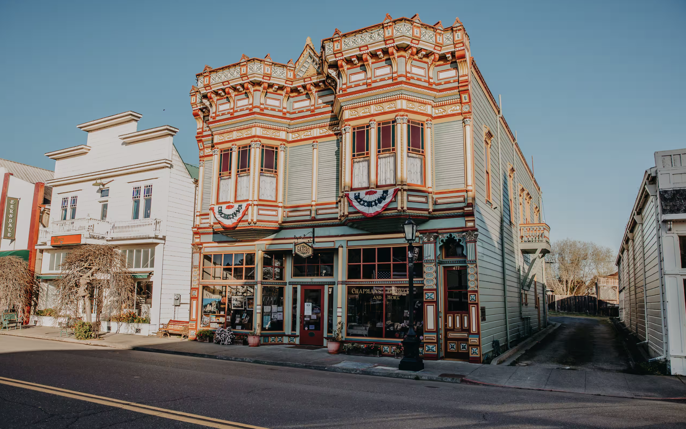 Ornately decorated historic storefronts line a sunlit street, with the central building displaying red, white, and blue bunting and signs for Gazebo.
