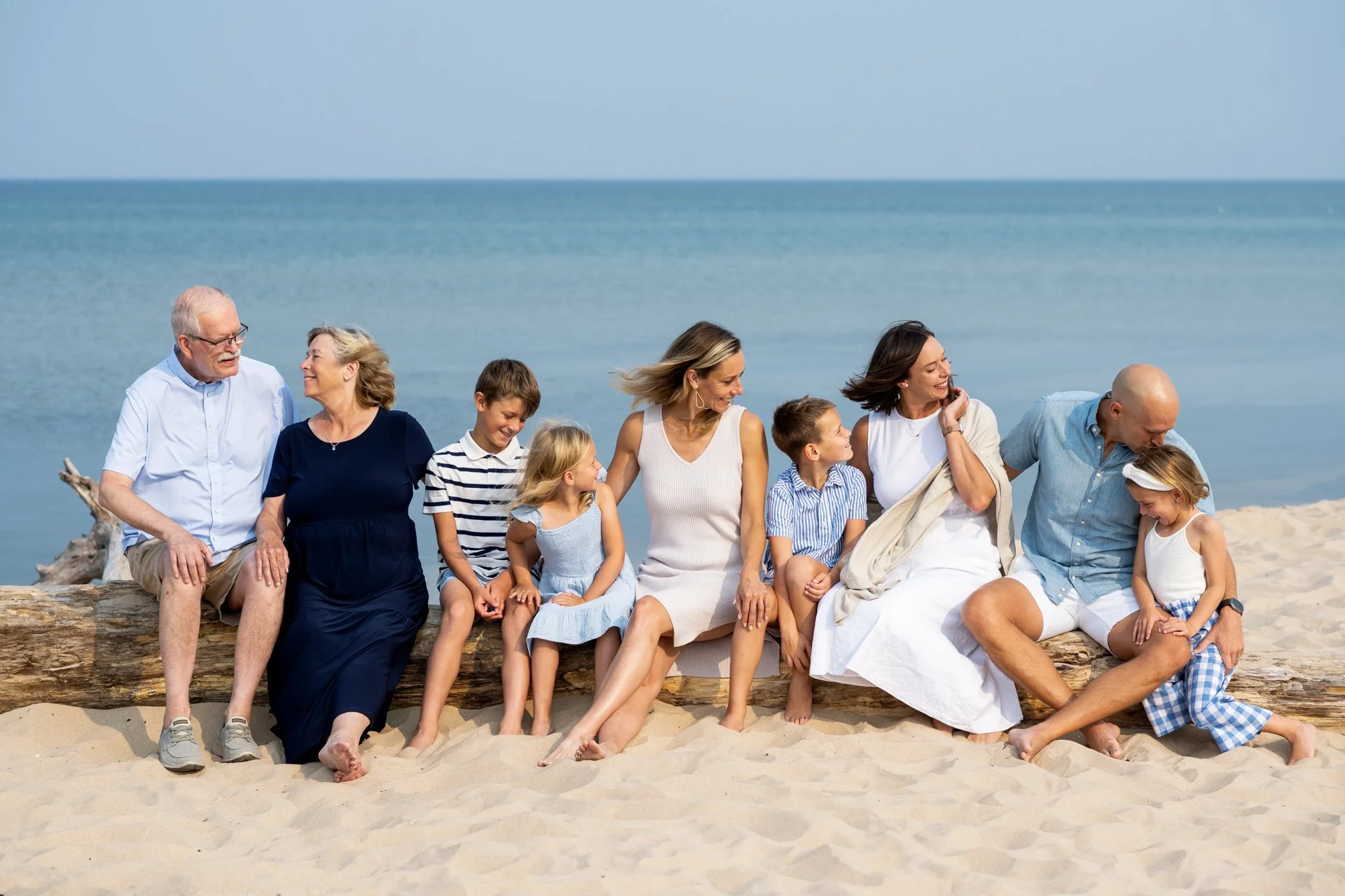 Family sitting on a beach log by the ocean, enjoying each other's company and smiling at each other.