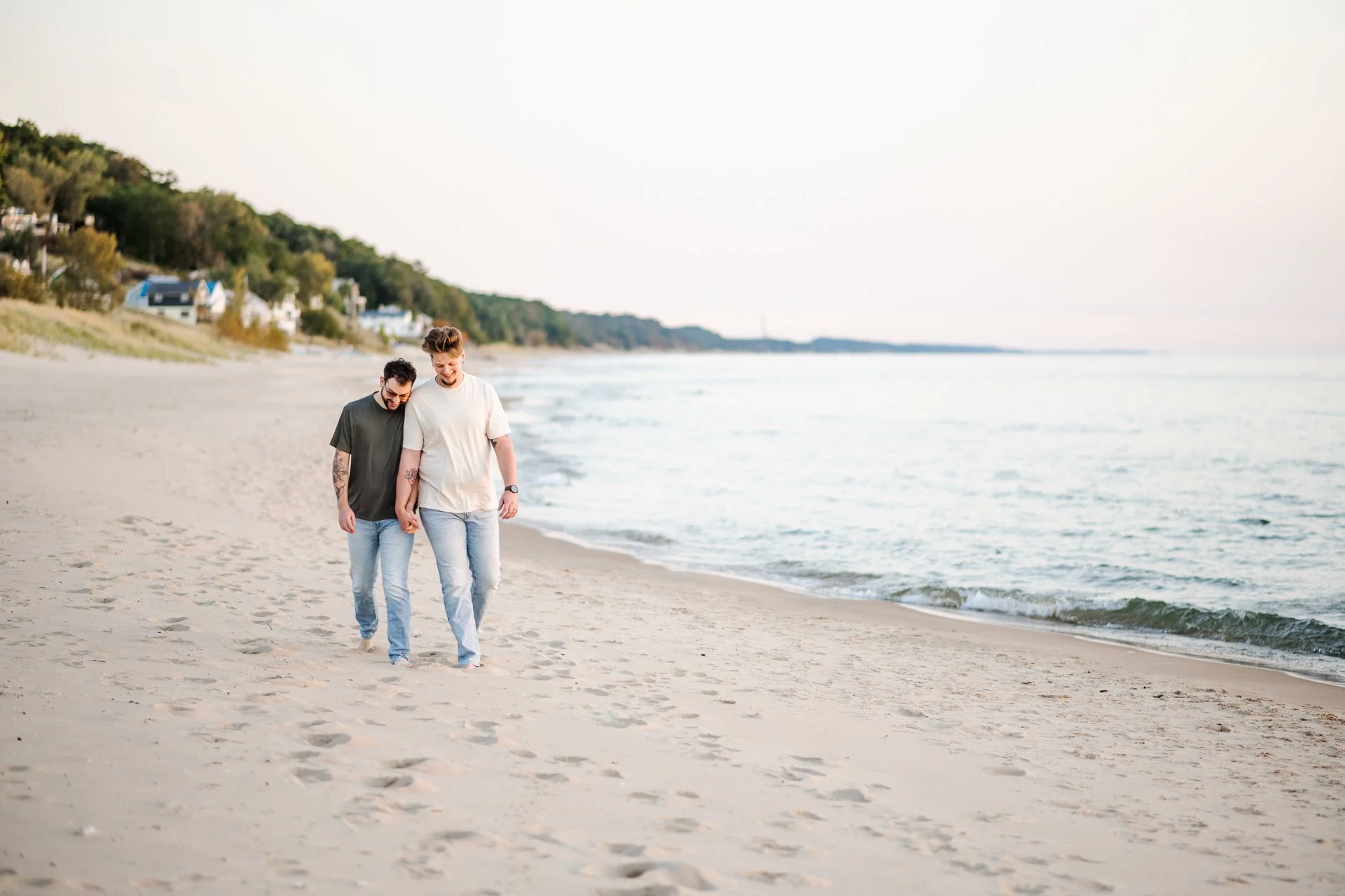 Two young men walk hand in hand along a sandy beach near the water with houses and trees in the background.