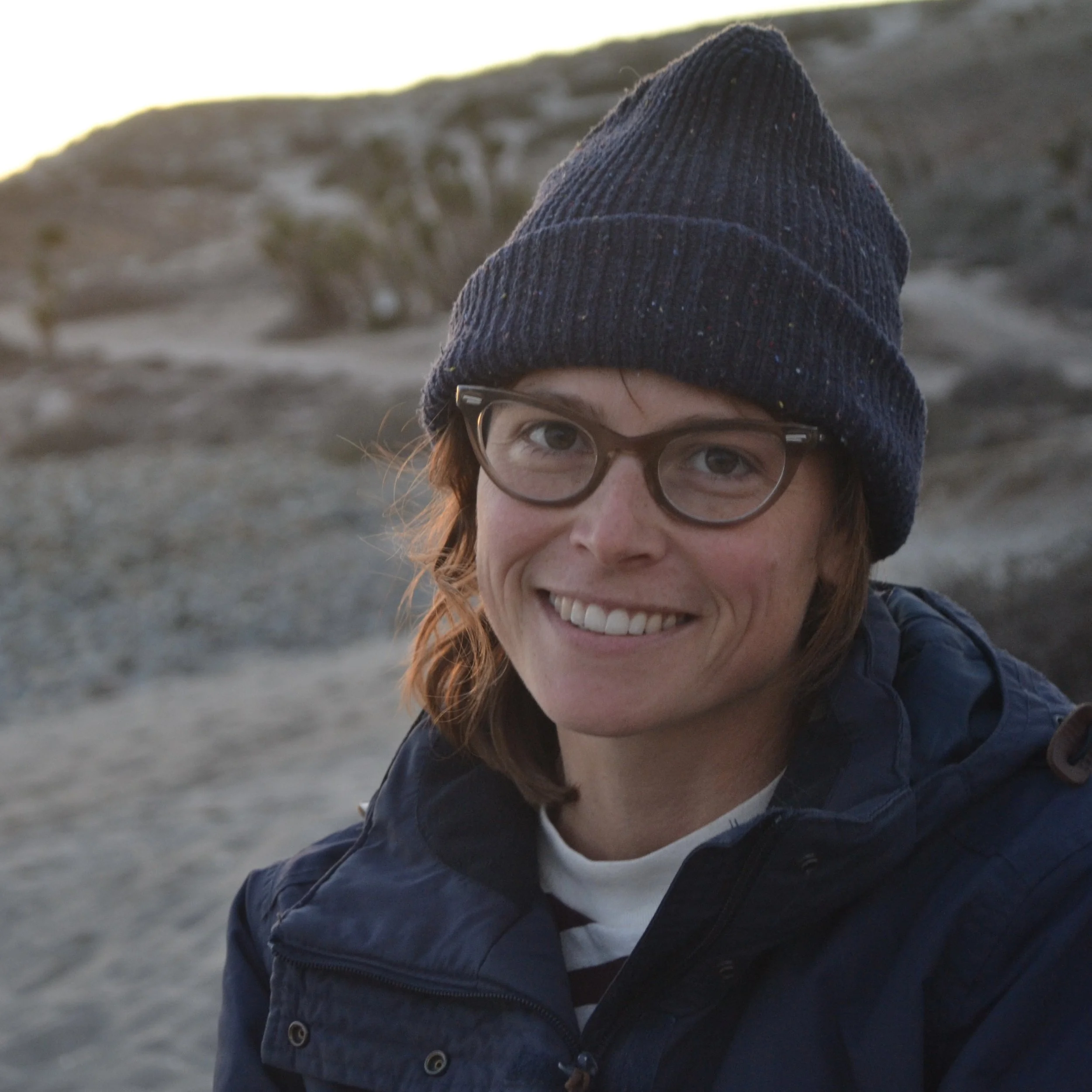 Portrait of smiling white woman with brown hair wearing brown glasses and a navy beanie on a sandy desert beach.
