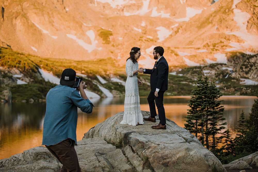 capturing couple with a film camera, bride and groom shake hands, high alpine lake elopement, mountain peak elopement, spring snow runoff, snow laiden peaks, national forest elopement