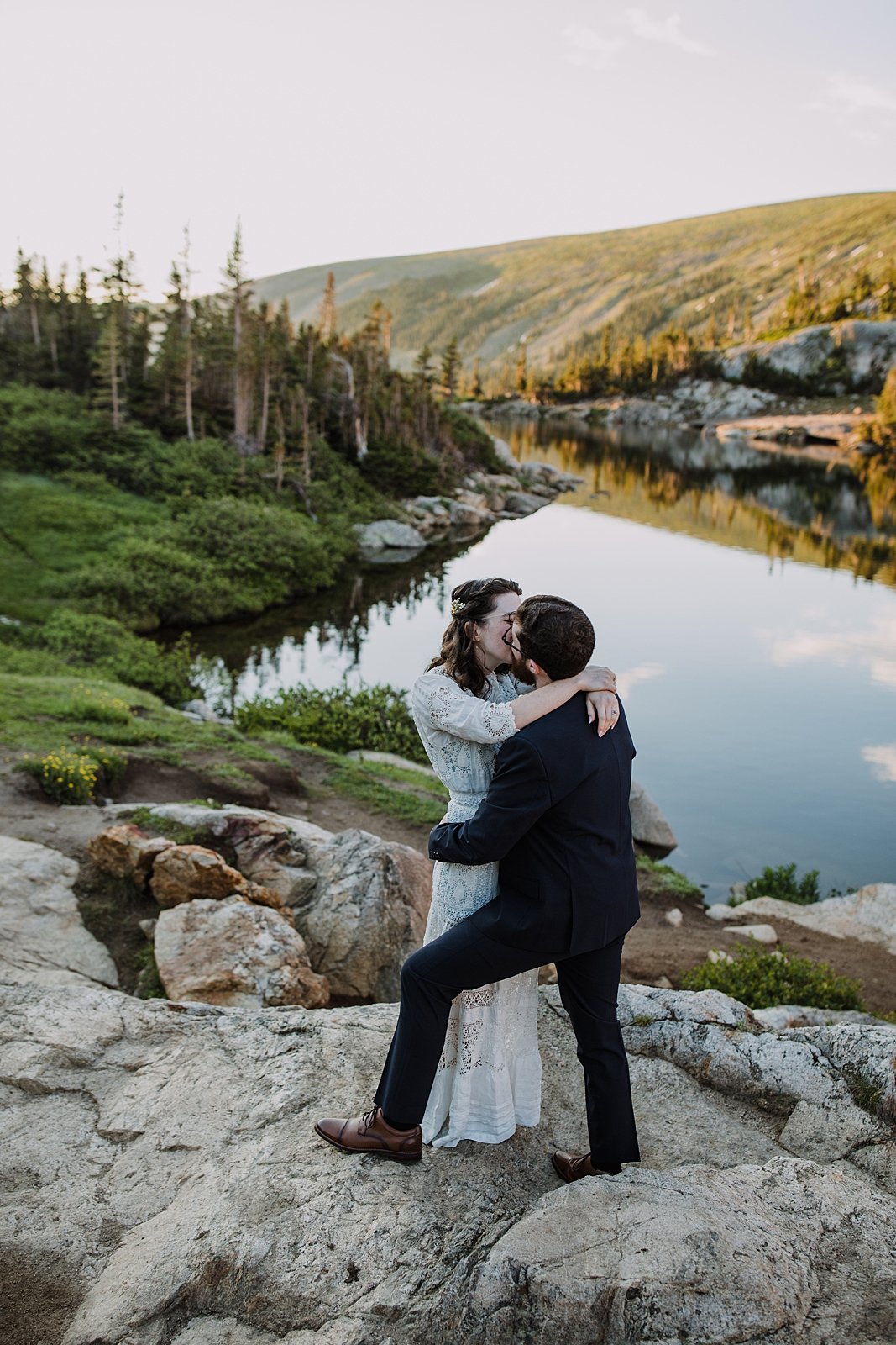 groom dipping bride for a kiss, reflection of pine trees on alpine lake, mitchell lake elopement, early morning elopement, antique bride and groom wedding attire, elopement hairstyle