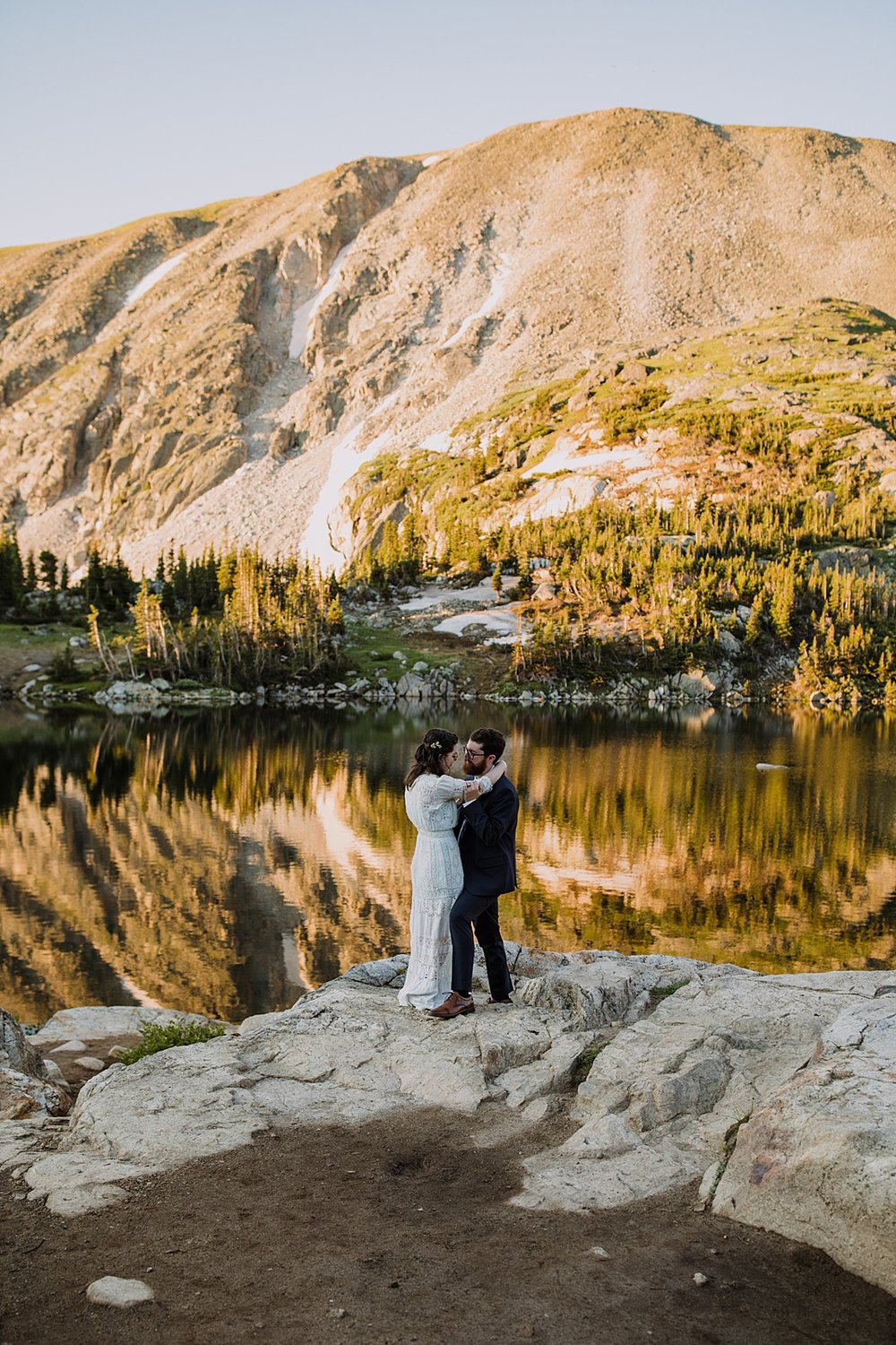 groom dipping bride for a kiss, reflection of pine trees on alpine lake, mitchell lake elopement, early morning elopement, antique bride and groom wedding attire, elopement hairstyle